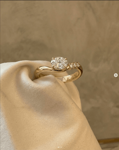 Yellow gold bypass engagement ring with round brilliant diamond and pavé accent stones on cream fabric